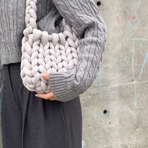 May include: A light gray, chunky knit shoulder bag with a braided strap. The bag is held in front of a person wearing a gray cable knit sweater and dark gray pants. The background is a concrete wall.