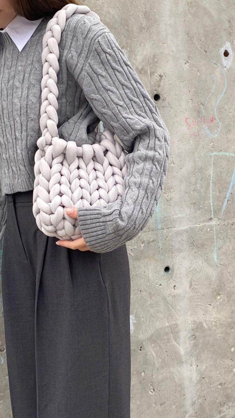 May include: A light gray, chunky knit shoulder bag with a braided strap. The bag is held in front of a person wearing a gray cable knit sweater and dark gray pants. The background is a concrete wall.