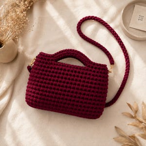 Puede incluir: Un bolso de ganchillo de color burdeos intenso con cuerpo rectangular, asa superior gruesa integrada y una correa larga trenzada a juego sujeta con cierres de metal dorado. El bolso presenta un patrón de punto denso y texturizado.