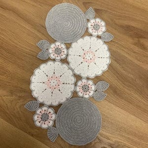 May include: A handmade crochet table runner featuring two solid grey circular mats, two large white floral motifs with pale pink centres, and three smaller white and grey floral accents with pale pink centres, all connected by grey crocheted leaves.