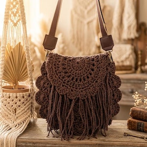 Puede incluir: Un bolso de hombro de ganchillo marrón con solapa festoneada y flecos. El bolso tiene una correa marrón y herrajes dorados. El bolso está sobre una superficie de madera.