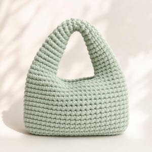 Puede incluir: Un bolso de mano color verde menta claro confeccionado con cordón grueso en un patrón de punto de ganchillo denso. El bolso presenta un diseño de asa integrada redondeada con una base sólida y estructurada y una textura uniforme.