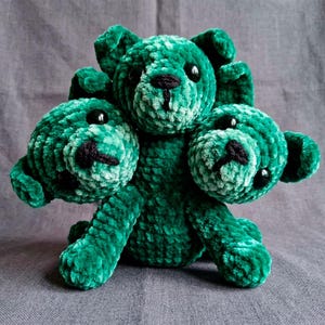 May include: A teal-coloured, three-headed plush bear. The toy has black button eyes and black stitching for the nose and mouth. The soft, textured fabric gives the bear a cuddly appearance. A handmade item.