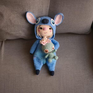 Può includere: Bambola all'uncinetto fatta a mano con una tuta blu con cappuccio e grandi orecchie. La bambola ha i capelli biondi, un fiocco rosa e tiene in mano una piccola creatura verde. I lineamenti della bambola sono dettagliati con accenti neri e rosa.