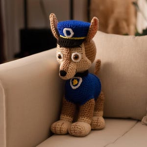 May include: A crocheted toy dog, resembling a cartoon character, wearing a blue police hat and uniform. The dog is tan with brown eyes and a badge. It is positioned on a beige sofa.