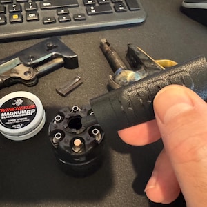 Puede incluir: Un surtido de piezas de armas de fuego antiguas, incluyendo un cilindro de revólver, una lata de pólvora negra etiquetada "Winchester Magnum BP Black Powder", y una parte de una pistola. Vista superior.