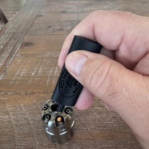 May include: A black cylinder with the text "REED RANCH BLACK POWDER" is held over a silver-colored revolver cylinder. The revolver cylinder has six chambers. The image is taken on a wooden table.