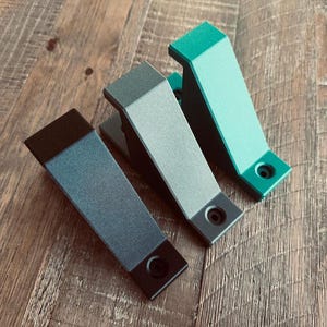 May include: Three metal cabinet pulls in shades of black, gray, and teal green are arranged on a wooden surface. Each pull has a rectangular body with a rounded top and a circular hole for mounting. The pulls are angled slightly.
