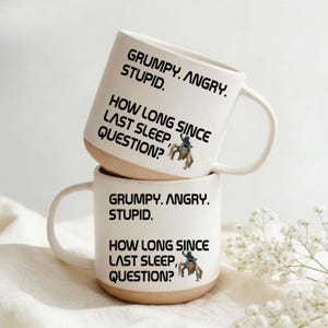 Könnte beinhalten: Zwei weiße Keramikbecher übereinander gestapelt. Jeder Becher hat einen beigefarbenen Boden und einen gebogenen Henkel. Die Becher tragen den Text "GRUMPY. ANGRY. STUPID." und "HOW LONG SINCE LAST SLEEP, QUESTION?" in Schwarz.