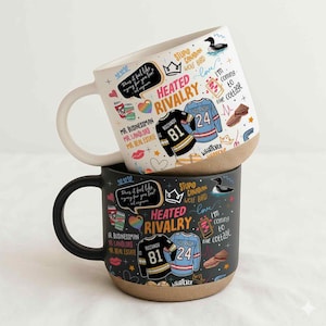 May include: Two ceramic mugs, one white and one black, stacked. Both mugs feature the text "Heated Rivalry" and various doodles, including hockey jerseys with the numbers 81 and 24. The white mug has a beige base and a white handle.