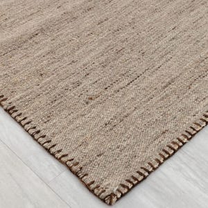 A close-up of a corner of the beige flatweave rug, highlighting the brown stitched edge and the woven texture.