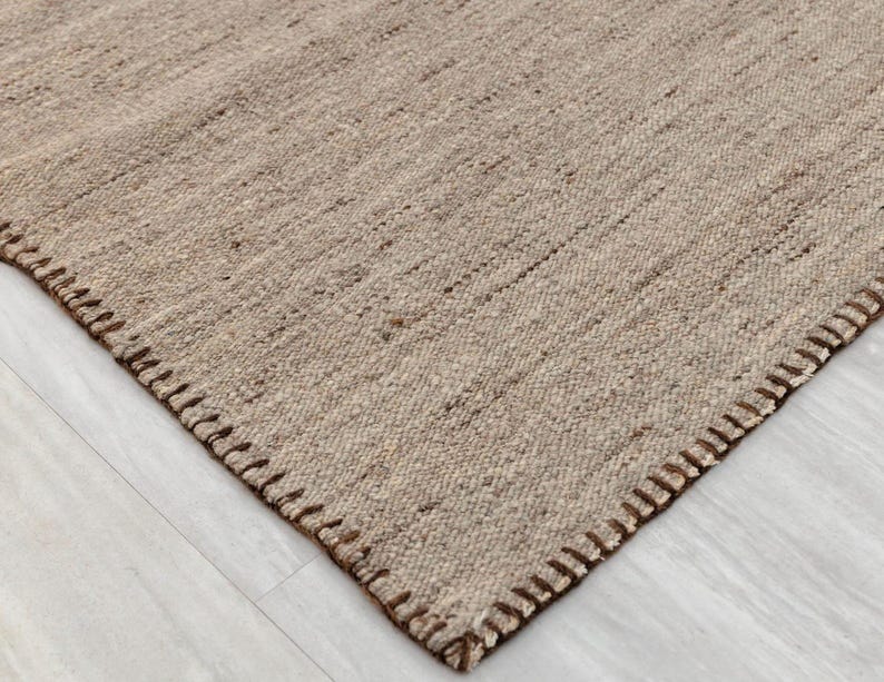 A close-up of a corner of the beige flatweave rug, highlighting the brown stitched edge and the woven texture.