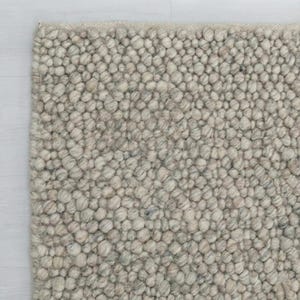 Puede incluir: Primer plano de una alfombra texturizada de tonos neutros. La superficie de la alfombra está cubierta de pequeños detalles redondeados y nudosos, creando un patrón táctil y visualmente interesante. La paleta de colores se compone principalmente de tonos grises claros y beige.