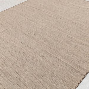 A full view of a rectangular beige flatweave rug with brown stitched edges, laid flat on a light-colored floor.