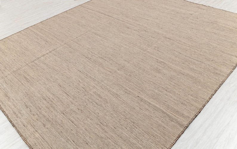 A full view of a rectangular beige flatweave rug with brown stitched edges, laid flat on a light-colored floor.