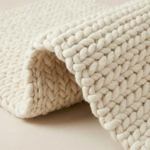 May include: Close-up of a cream-coloured, chunky knit blanket. The thick yarn is tightly woven, creating a textured surface. The blanket is folded over, revealing the inside of the knit. The overall look is cosy and warm.