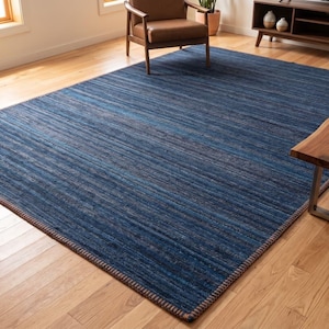 Puede incluir: Una alfombra azul rectangular grande con un patr&oacute;n de rayas. La alfombra est&aacute; hecha de un material tejido y tiene un borde marr&oacute;n. La alfombra est&aacute; colocada sobre un piso de madera en un sal&oacute;n.
