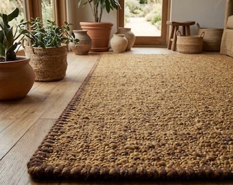 Puede incluir: Una alfombra rectangular con una superficie tejida gruesa y texturizada de color marrón dorado y un borde cosido de color marrón más oscuro. La alfombra está colocada sobre un suelo de madera clara, extendiéndose desde el frente hacia el fondo.