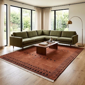 May include: An L-shaped sectional sofa upholstered in olive green fabric with wooden legs, positioned on a large rectangular area rug in burnt orange with black geometric border patterns and fringe, accompanied by a low wooden coffee table.