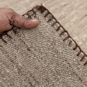 A hand holds up a corner of a beige flatweave rug, showing the brown stitched edge and the texture of the woven fibers.