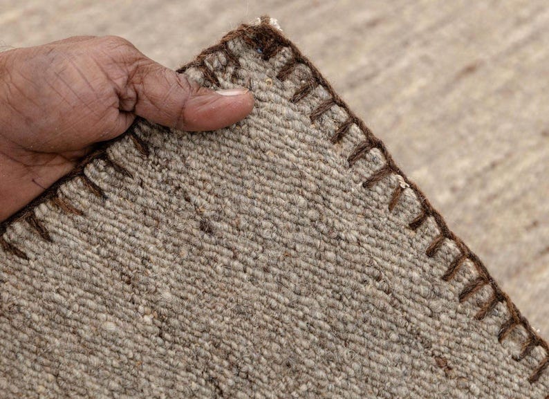 A hand holds up a corner of a beige flatweave rug, showing the brown stitched edge and the texture of the woven fibers.