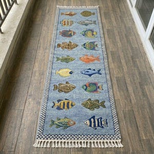 May include: A rectangular blue rug with a repeating pattern of colorful fish. The rug has a white fringe on the short ends and a patterned border. The fish are in various colors, including yellow, orange, and blue.