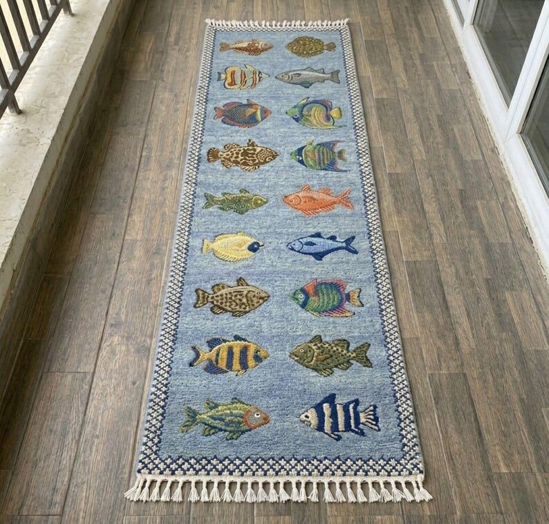 May include: A rectangular blue rug with a repeating pattern of colorful fish. The rug has a white fringe on the short ends and a patterned border. The fish are in various colors, including yellow, orange, and blue.