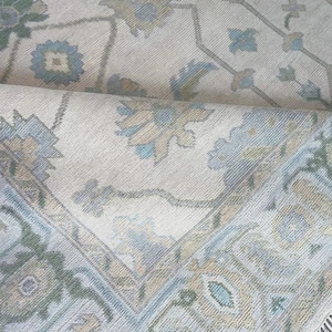May include: A close-up of a beige rug with a floral pattern in shades of blue, green, and tan. The rug has a rolled edge, revealing the intricate design and fringe detail. The rug's design features stylized floral and geometric elements.