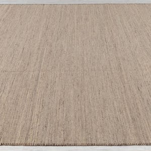 An overhead, slightly angled view of a rectangular beige flatweave rug with brown stitched edges, laid on a light floor.