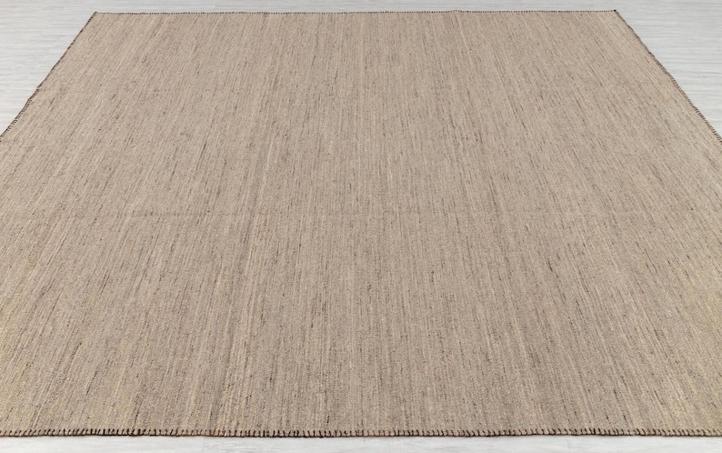 An overhead, slightly angled view of a rectangular beige flatweave rug with brown stitched edges, laid on a light floor.