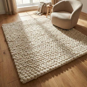 May include: A rectangular area rug featuring a thick, chunky knit texture made of cream-colored wool yarn. The rug has a dense, pebble-like surface pattern created by large, interconnected loops of soft material, providing a uniform and tactile appearance.