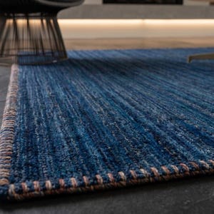 Puede incluir: Una alfombra azul rectangular con una superficie texturizada y un borde contrastante. La paleta de colores de la alfombra incluye tonos de azul y un borde marr&oacute;n. La alfombra est&aacute; colocada sobre un suelo de baldosas gris oscuro, con una silla y un jarr&oacute;n en el fondo.