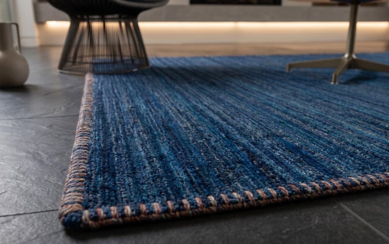 Puede incluir: Una alfombra azul rectangular con una superficie texturizada y un borde contrastante. La paleta de colores de la alfombra incluye tonos de azul y un borde marr&oacute;n. La alfombra est&aacute; colocada sobre un suelo de baldosas gris oscuro, con una silla y un jarr&oacute;n en el fondo.