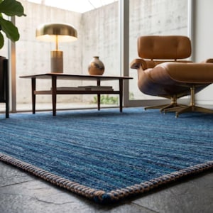 Puede incluir: Una alfombra azul rectangular grande con una superficie texturizada. La alfombra tiene un borde tejido en tonos marr&oacute;n y crema. La alfombra est&aacute; en un sal&oacute;n moderno con una silla de cuero, una mesa de centro y una planta en maceta.