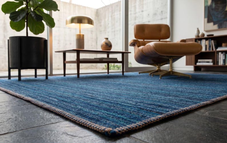Puede incluir: Una alfombra azul rectangular grande con una superficie texturizada. La alfombra tiene un borde tejido en tonos marr&oacute;n y crema. La alfombra est&aacute; en un sal&oacute;n moderno con una silla de cuero, una mesa de centro y una planta en maceta.