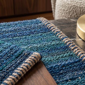 Puede incluir: Una alfombra tejida azul y turquesa con un borde de fibra natural. La alfombra tiene un aspecto texturizado y est&aacute; colocada sobre una superficie oscura. Una base de l&aacute;mpara dorada es visible en el fondo.