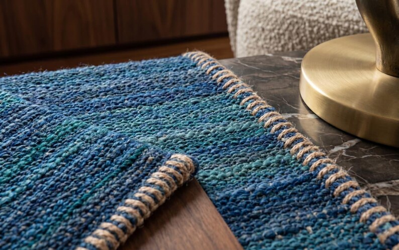 Puede incluir: Una alfombra tejida azul y turquesa con un borde de fibra natural. La alfombra tiene un aspecto texturizado y est&aacute; colocada sobre una superficie oscura. Una base de l&aacute;mpara dorada es visible en el fondo.