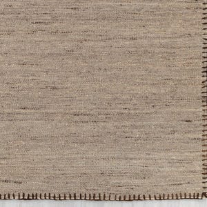 A close-up view of the beige flatweave rug, showing the tight weave and the brown stitched edge.