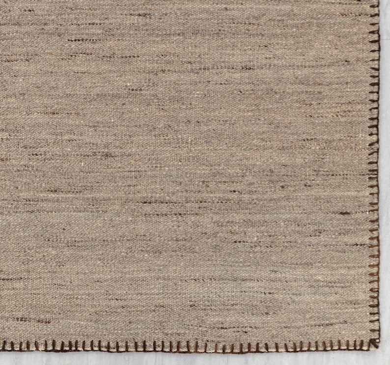 A close-up view of the beige flatweave rug, showing the tight weave and the brown stitched edge.