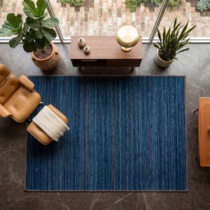 Puede incluir: Vista a&eacute;rea de un sal&oacute;n moderno. Una alfombra azul a rayas es el centro, con un sill&oacute;n y un otom&aacute;n de cuero marr&oacute;n a la izquierda. Una mesa auxiliar de madera y una peque&ntilde;a mesa de madera est&aacute;n a ambos lados de la alfombra. Tambi&eacute;n se ven plantas y una l&aacute;mpara.