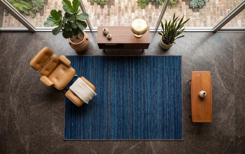 Puede incluir: Vista a&eacute;rea de un sal&oacute;n moderno. Una alfombra azul a rayas es el centro, con un sill&oacute;n y un otom&aacute;n de cuero marr&oacute;n a la izquierda. Una mesa auxiliar de madera y una peque&ntilde;a mesa de madera est&aacute;n a ambos lados de la alfombra. Tambi&eacute;n se ven plantas y una l&aacute;mpara.