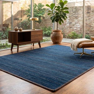 Puede incluir: Una alfombra azul rectangular grande con una superficie texturizada domina una sala de estar. La alfombra est&aacute; bordeada con un borde m&aacute;s oscuro y se asienta sobre un piso de madera. La habitaci&oacute;n presenta una est&eacute;tica moderna de mediados de siglo con una silla de cuero y muebles de madera.