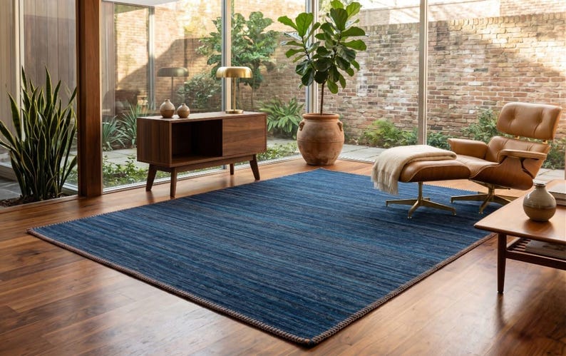 Puede incluir: Una alfombra azul rectangular grande con una superficie texturizada domina una sala de estar. La alfombra est&aacute; bordeada con un borde m&aacute;s oscuro y se asienta sobre un piso de madera. La habitaci&oacute;n presenta una est&eacute;tica moderna de mediados de siglo con una silla de cuero y muebles de madera.