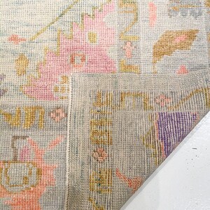 May include: A section of a light blue rug with a floral pattern in pink, yellow, and purple. The rug's design includes geometric shapes and a border. The rug is folded over, revealing the underside with a detailed border.