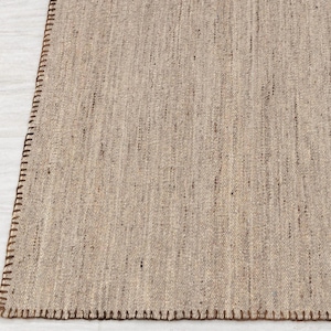 A corner view of the beige flatweave rug, showing the brown stitched edge and the texture of the woven surface.