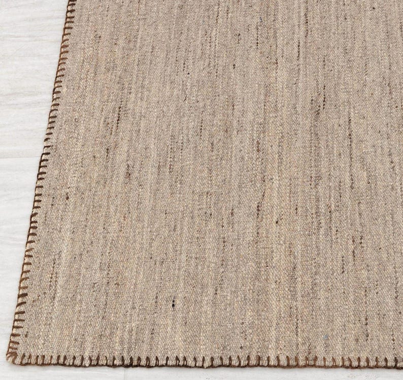 A corner view of the beige flatweave rug, showing the brown stitched edge and the texture of the woven surface.