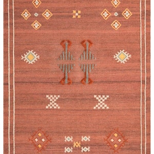 May include: A rectangular area rug in a deep terracotta red hue featuring a white border frame and fringe on the top and bottom edges. The rug is decorated with various geometric patterns in white, yellow, and blue, including diamonds and crosses.