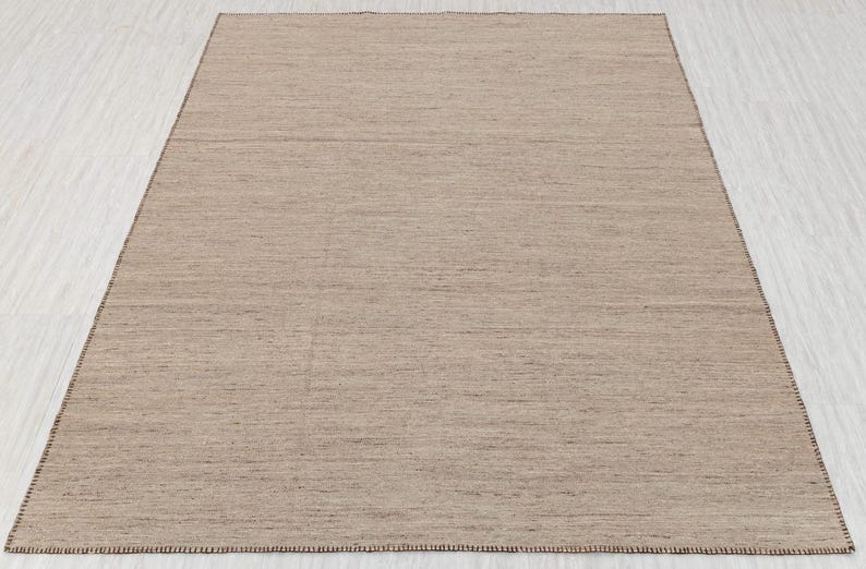A full, straight-on view of a rectangular beige flatweave rug with brown stitched edges, laid on a light-colored floor.
