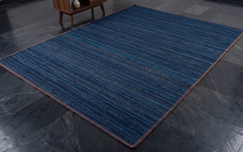 Puede incluir: Una alfombra rectangular azul con un patr&oacute;n de rayas. La alfombra es de color azul oscuro con acentos azules claros y marrones. Los bordes est&aacute;n acabados con un borde marr&oacute;n. La alfombra est&aacute; colocada sobre un suelo de baldosas gris oscuro.