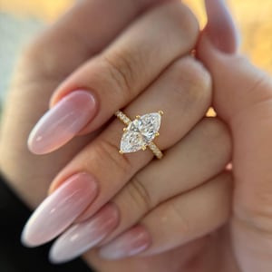 Op de afbeelding: Een gouden ring met een grote marquise-geslepen middensteen die door vier klauwen wordt vastgehouden, met een dunne band die aan de zijkanten is geaccentueerd met kleine pavé-gezette steentjes. Het metaal heeft een gepolijste gouden afwerking.